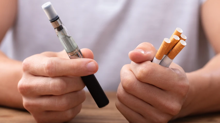 Man Holding Vape And Tobacco Cigarette Over Desk